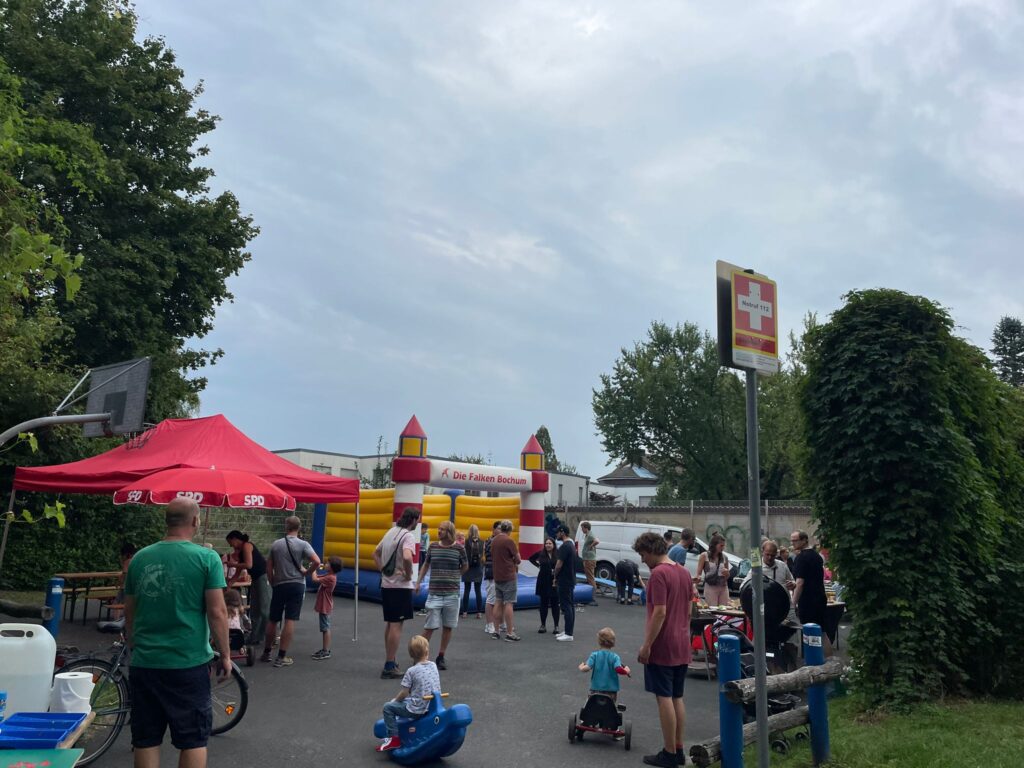 Mehrere Personen und Kinder stehen vor einem Sonnenschirm und einer Hüpfburg. 